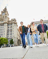 Vier Freunde erkunden eine lokale Stadt, während sie entlang der Uferpromenade spazieren gehen, mit historischen Gebäuden im Hintergrund.