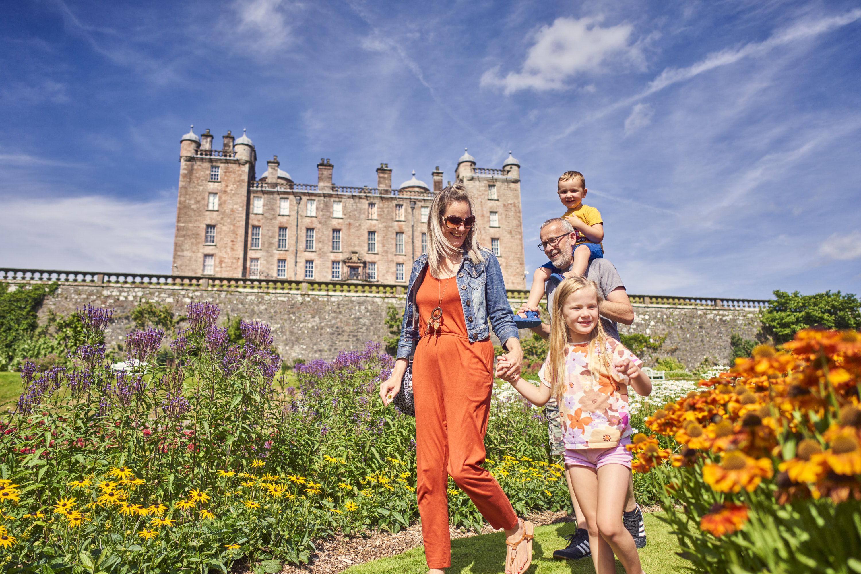 Family walking through colorful gardens in front of a historic stone castle under a blue sky, enjoying a sunny day outdoors.