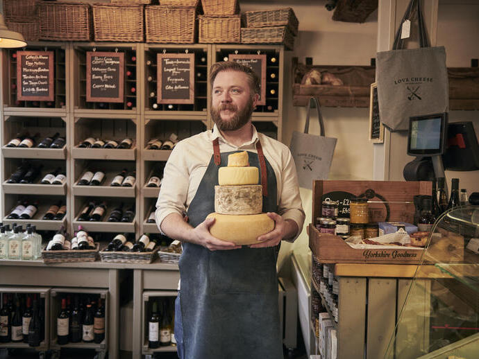 Man wearing blue apron holding a stack of cheeses in a cheese and wine shop