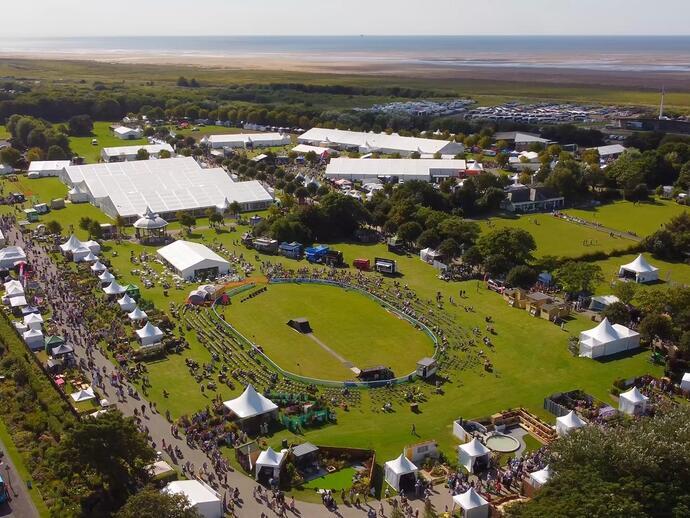 Aerial view of a large outdoor festival with crowds, tents, exhibition stands, event rings, lawns, and coastline in the background.