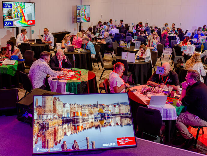 A conference room with round tables, people networking, laptops open, and a screen showing an image of Edinburgh, Scotland. Professional event atmosphere.