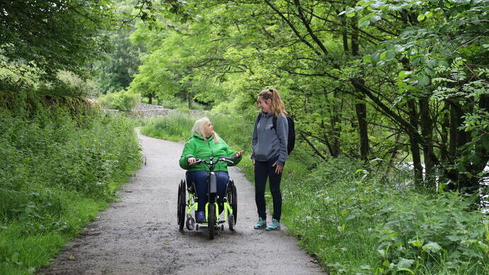 Zwei Frauen, eine auf einem Rollstuhlfahrrad, auf einem von Bäumen umgebenen Weg