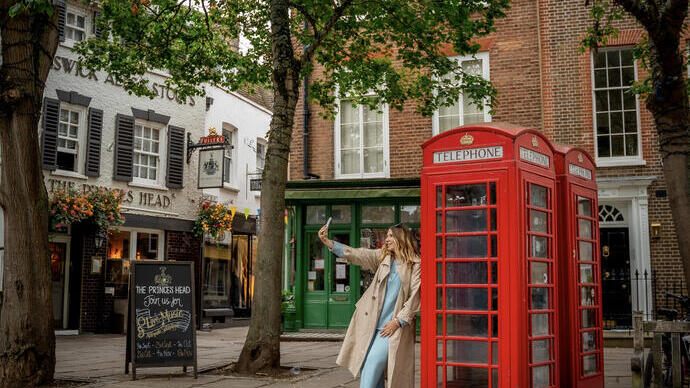 A woman leaning against a red telephone box and takes a selfie