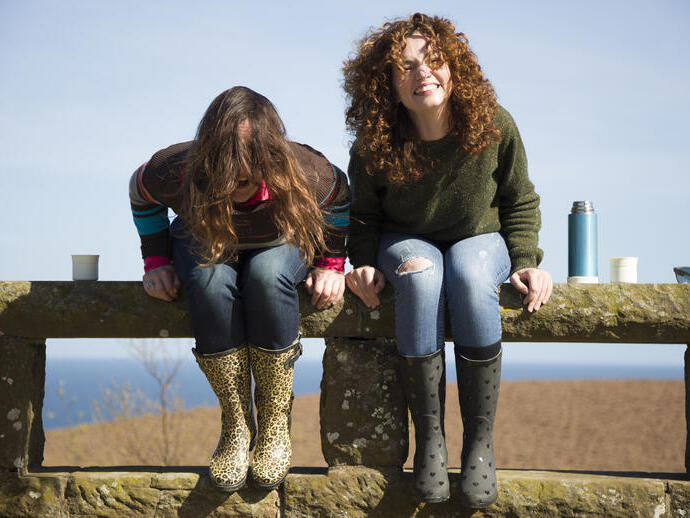 Two women laughing, sat on a stone fence