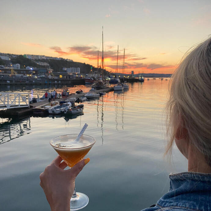 Eine Frau mit einem Cocktail in der Hand und Blick auf einen Hafen in der Ferne bei Sonnenuntergang