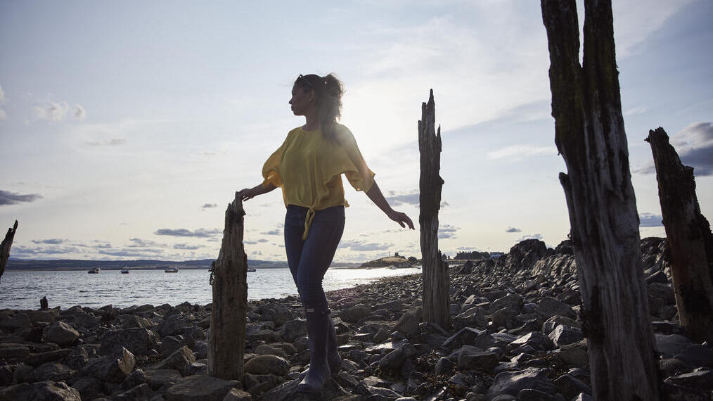 Woman walking on large stones in between old wooden stakes by the sea