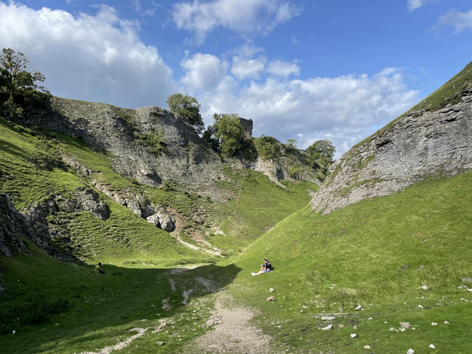 A valley on a bright summer's day, with a couple resting on the grass. A Castle is visible through the trees on the rocky hillside.