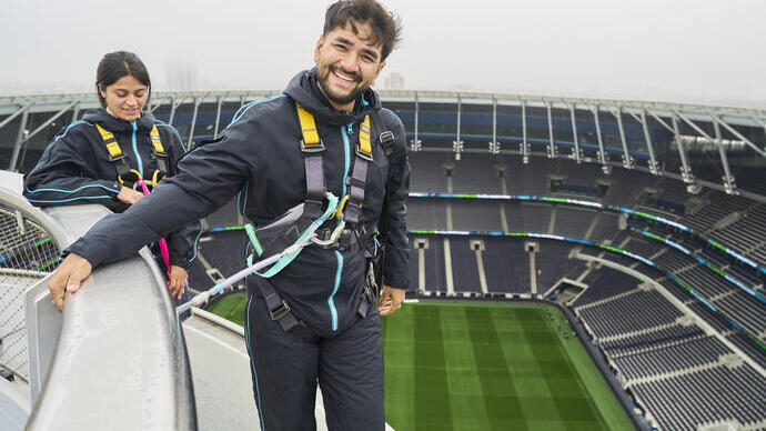 A man and woman wearing safety equipment stand on a platform above a football stadium