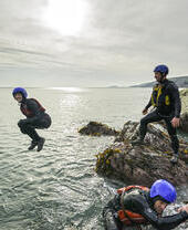 People in wetsuits and helmets coasteering by the sea, jumping and climbing on rocks under a cloudy sky, with scenic coastline in the background.
