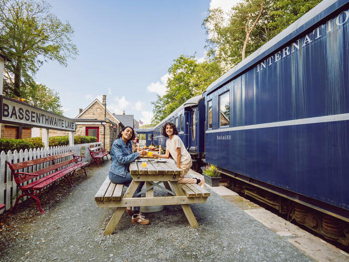 Two people dining at a picnic table on a historic train station platform with a blue train and Bassenthwaite Lake station sign visible.