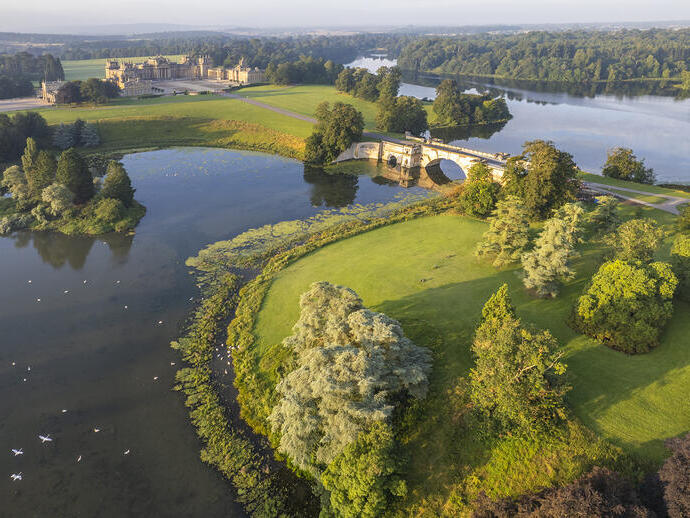 Vista aérea de un palacio del siglo XVIII, rodeado de jardines, árboles y un lago atravesado por un puente.