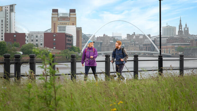 A woman and a man walk along a riverside with iconic buildings and a bridge beyond