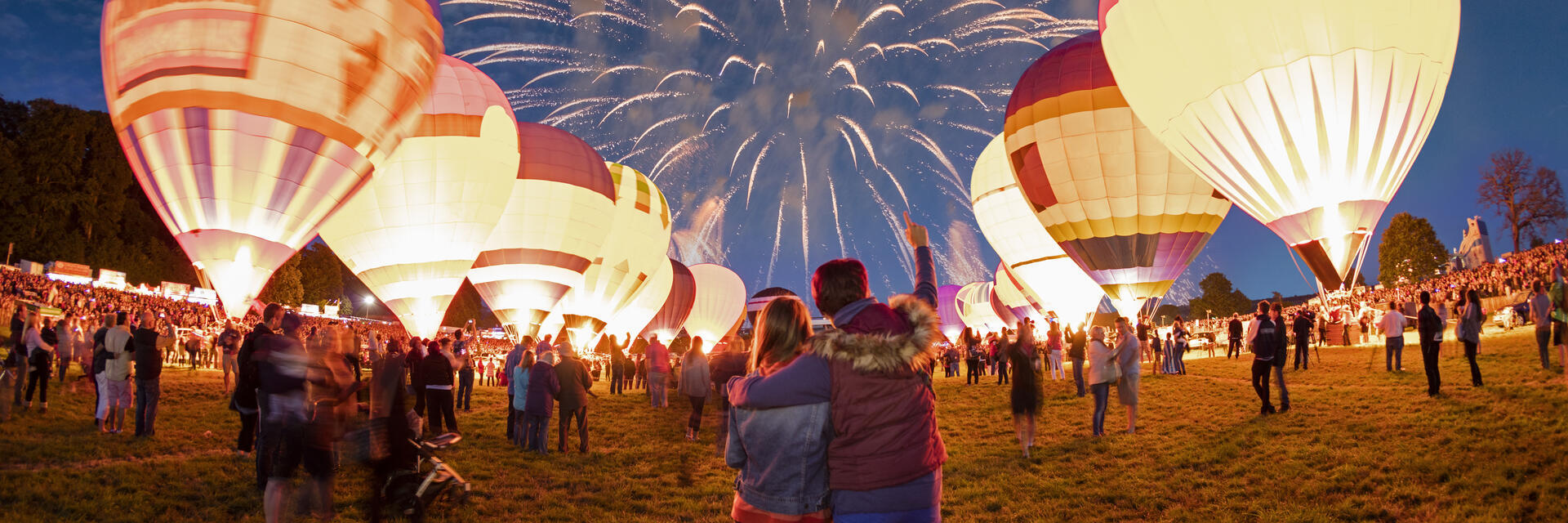 Pareja contemplando globos aerostáticos en tierra y fuegos artificiales en el cielo nocturno