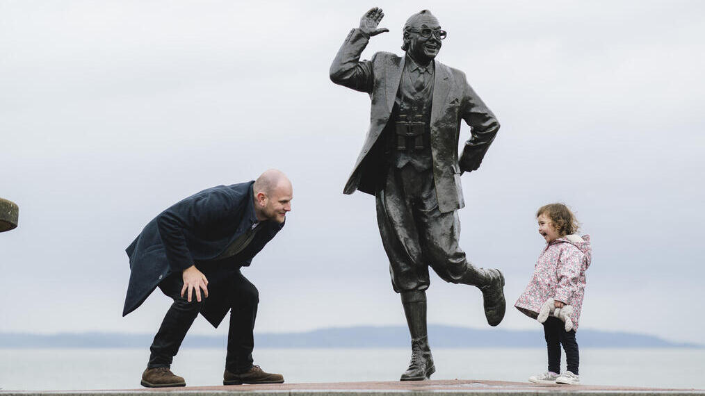A person and child interact playfully beside a statue on a seafront promenade, with the sea and distant hills in the background.