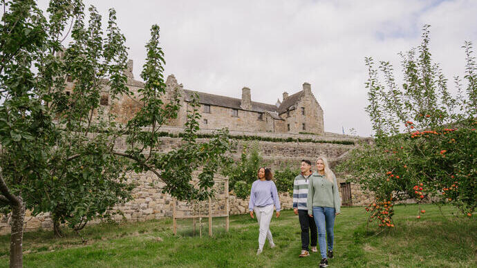Three people walk through an orchard in front of a historic stone building with cloudy skies above.