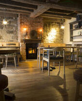 Cozy rustic room with wooden floors, exposed beams, brick fireplace, wooden tables and chairs, and decorative shelves.