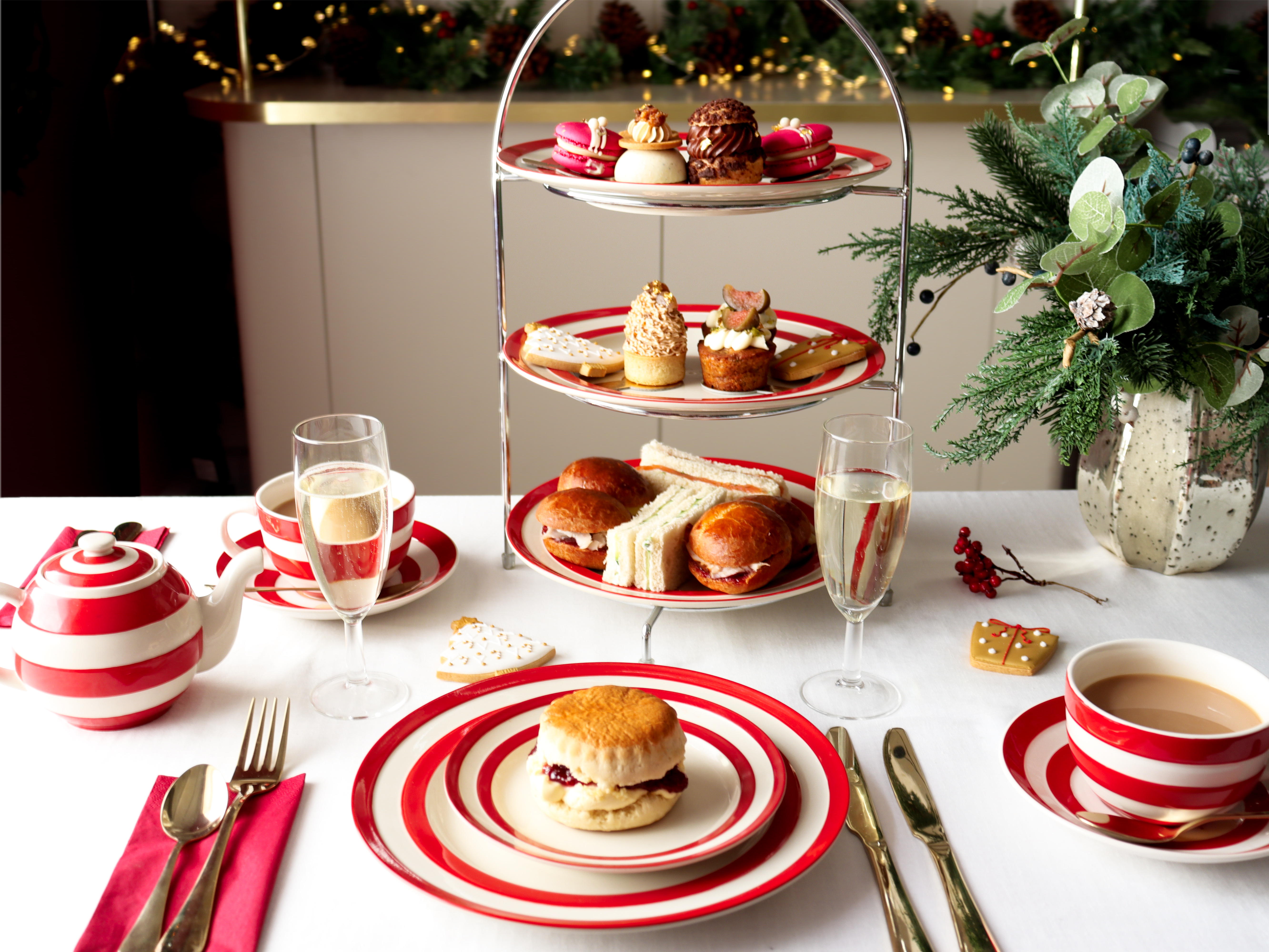 Mesa servida para té de la tarde con dulces, sándwiches, té, copas, vajilla rayada roja y blanca y decoraciones navideñas al fondo.