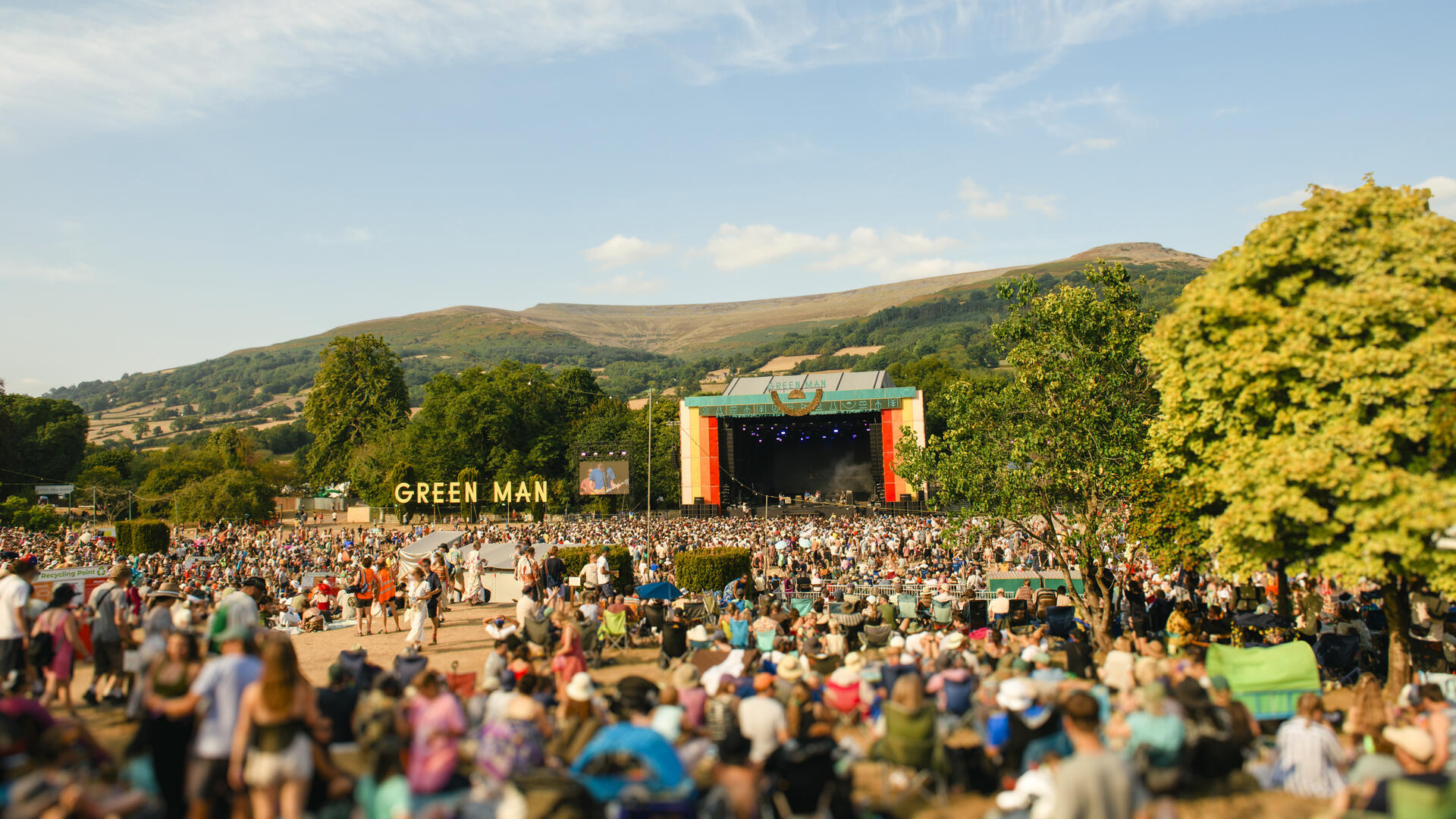 Large outdoor music festival crowd watching a stage, with mountains and trees in the background under a blue sky.