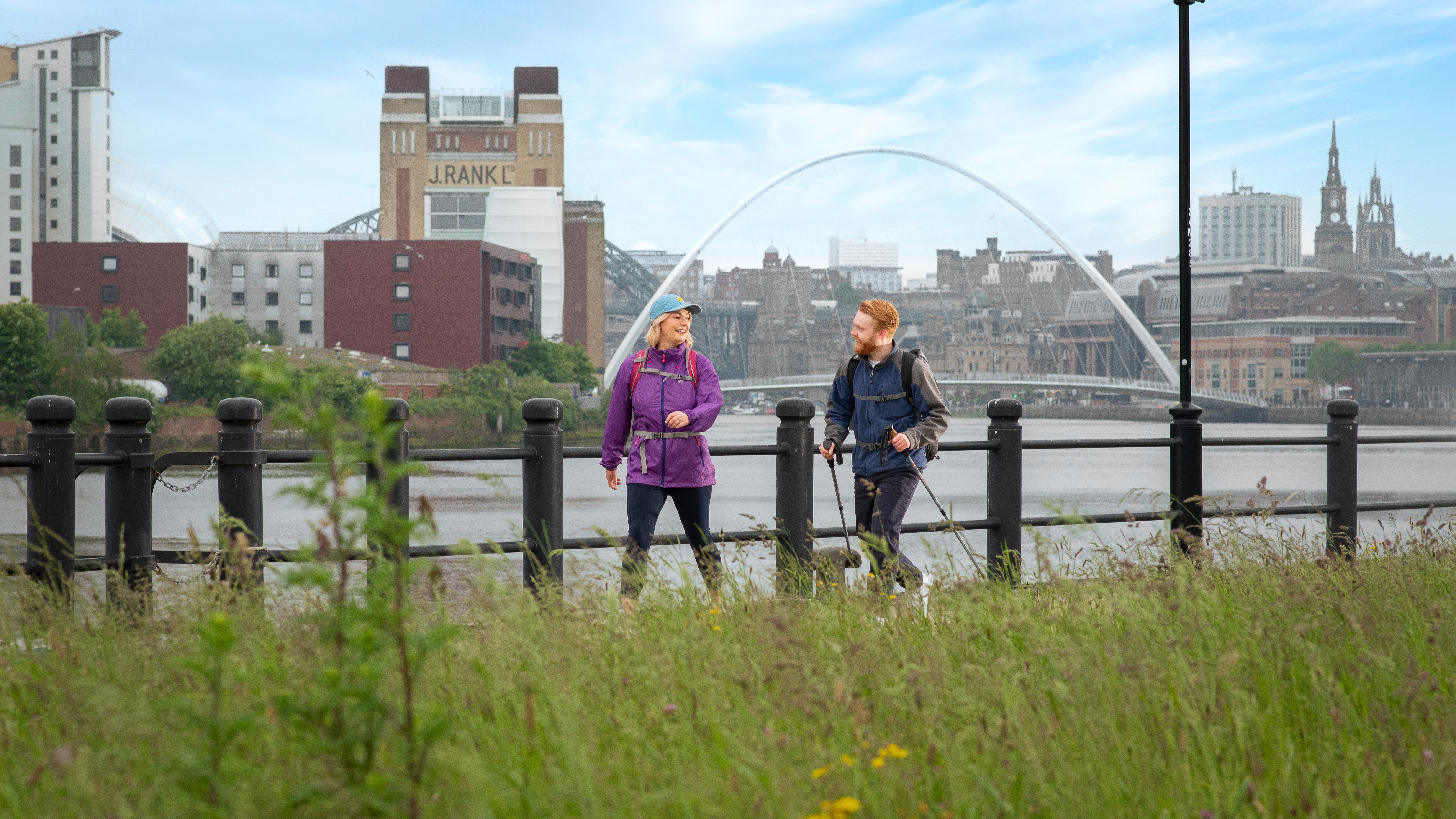 A woman and a man walk along a riverside with iconic buildings and a bridge beyond
