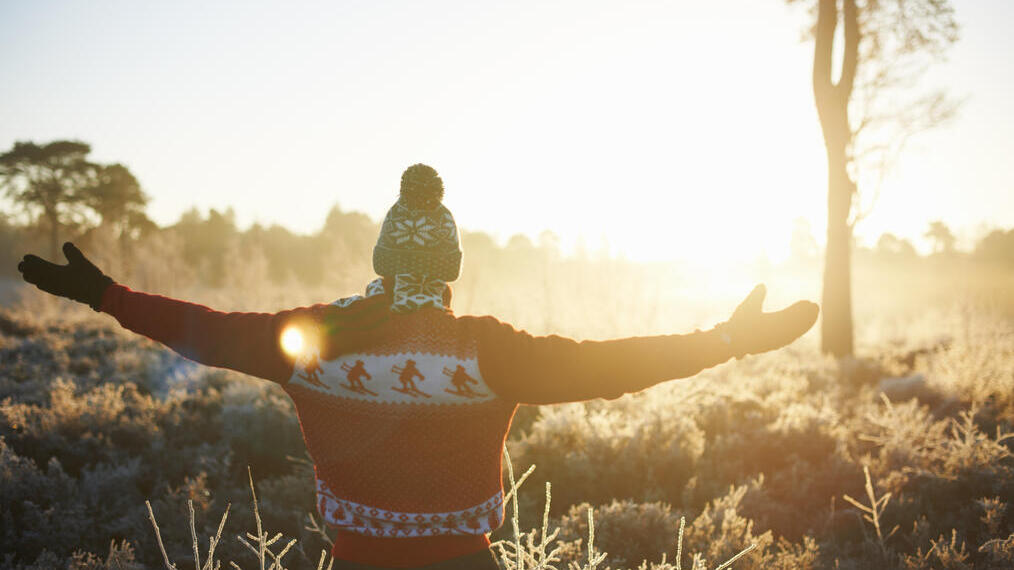Person in a festive sweater and hat stands in a frosty field with arms outstretched, facing the sunrise.