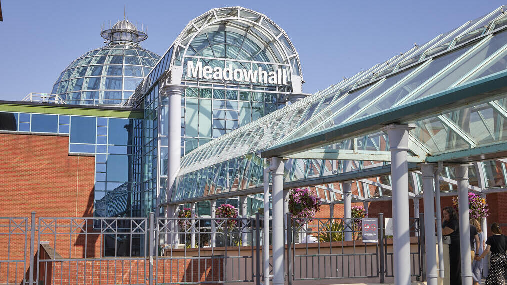 Modern glass entrance of Meadowhall shopping centre with dome, walkway, brick walls, and flower baskets under a clear blue sky.