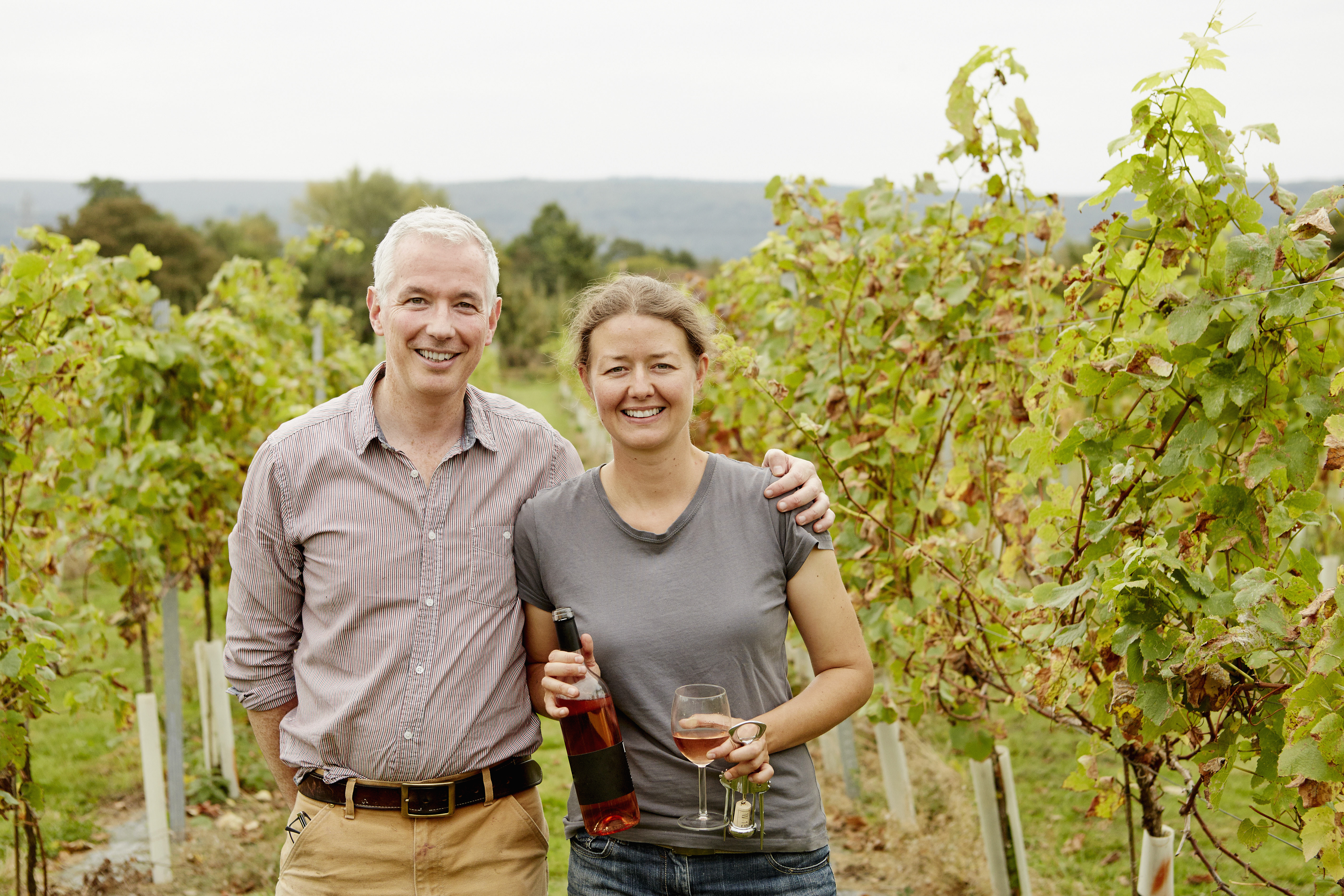 Couple standing in a vineyard holding wine and glasses.