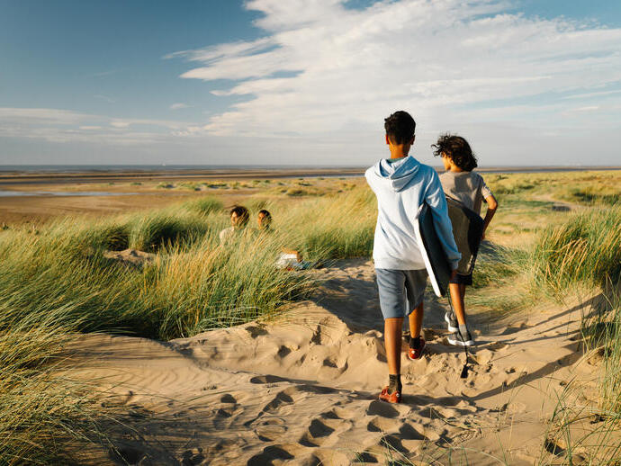 Two people walk with boards along sandy dunes to a wide beach under a blue sky, with two others sitting among the grass.