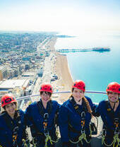 Group of friends at the top of i360 pod in climbing gear with coastline of Brighton. below