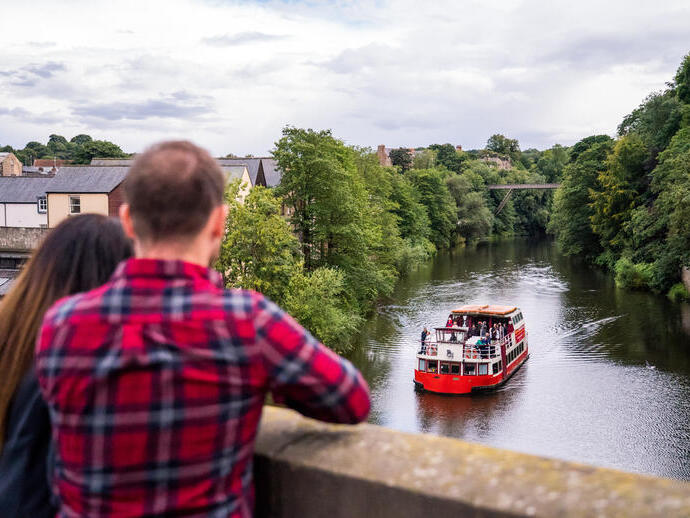 Zwei Personen stehen auf einer Brücke und beobachten ein Ausflugsboot, das auf einem Fluss in Durham fährt