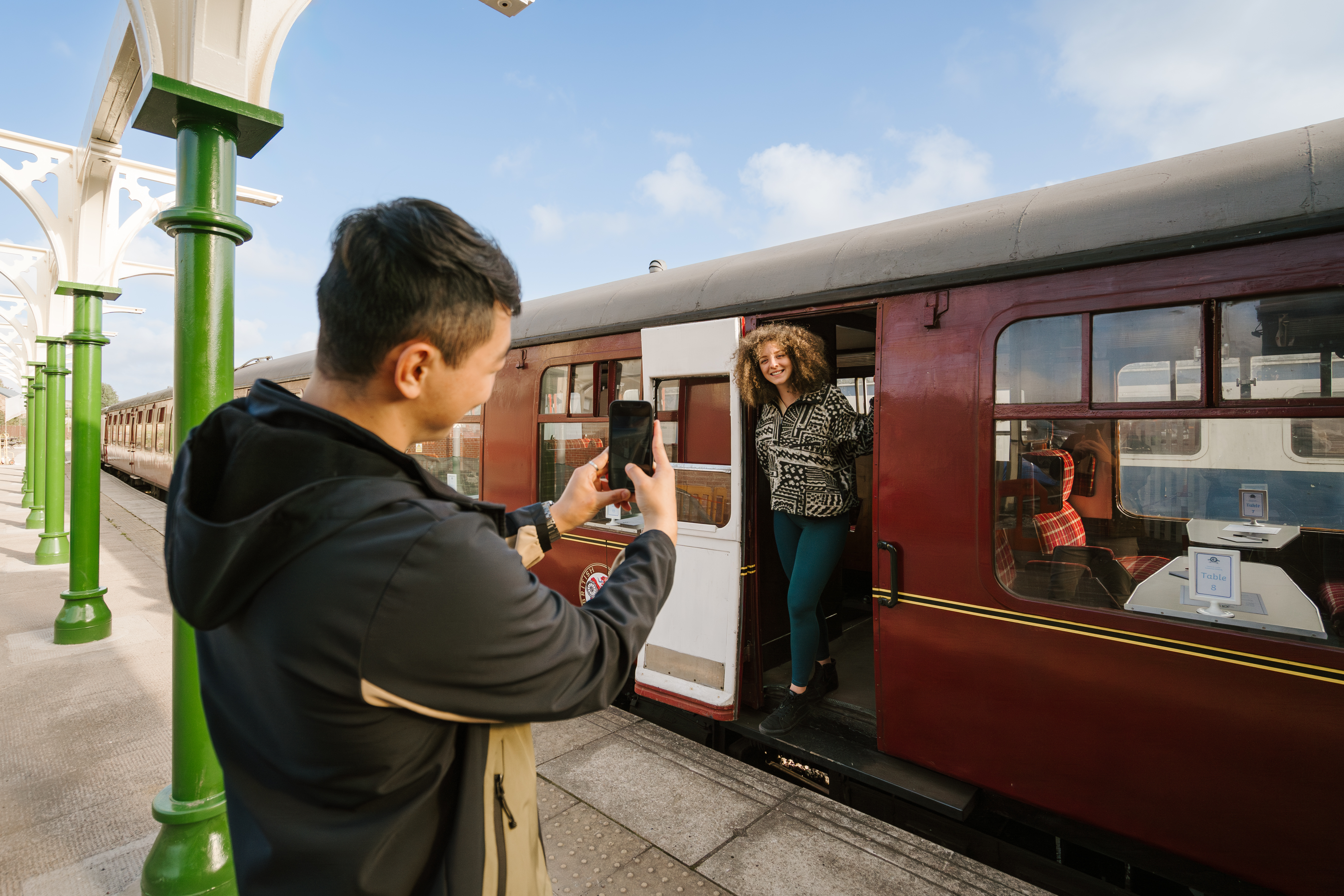 Un hombre tomando una foto de una mujer de pie junto a la puerta de un tren en el andén de una estación.