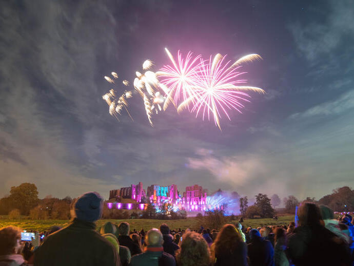 Fireworks display over a ruined castle watched by a crowd of people in the evening