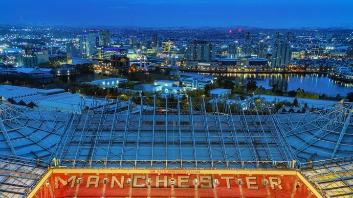 Aerial view of Old Trafford stadium with 'Manchester United' seats, city skyline and river at dusk, with illuminated buildings.