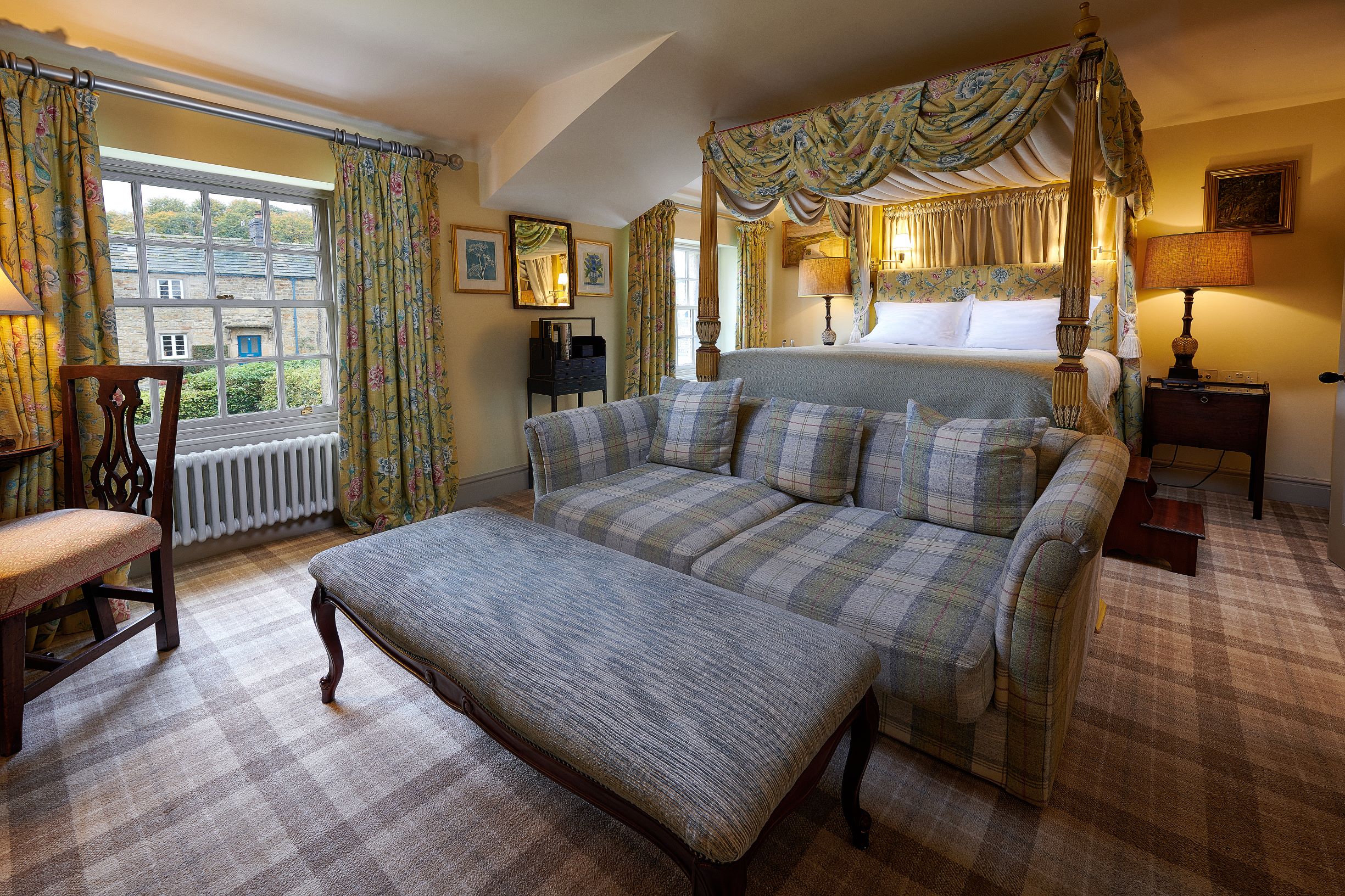 Bedroom at the Devonshire Arms at Pilsley, Derbyshire with a grey tartan upholstered sofa and large foot stool in the foreground, matching tartan carpet and floral curtains and matching draped four-poster bed in the background.