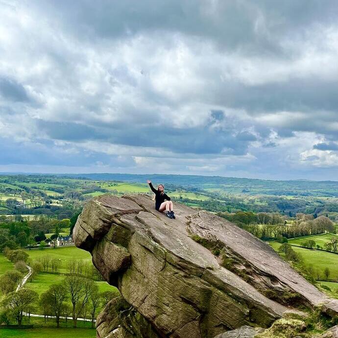Person sits on large rocky outcrop with scenic countryside and dramatic cloudy sky in the background.