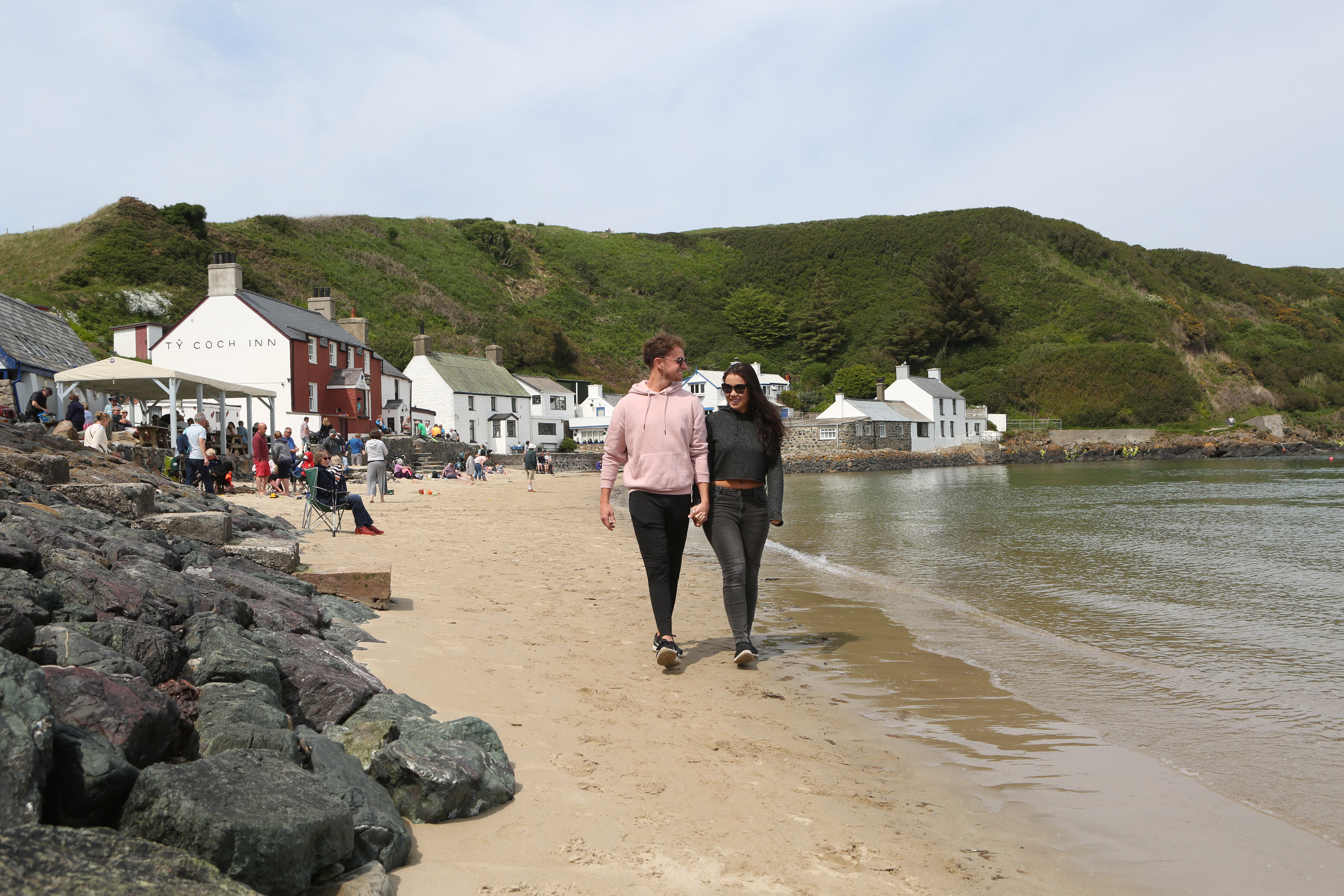 Two people walking along a sandy beach with the ocean to one side and pubs and shops on the other side.