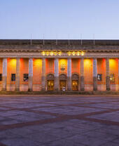 Large neoclassical building with columns, illuminated at dusk, facing an open square. Decorative lights accent the facade.