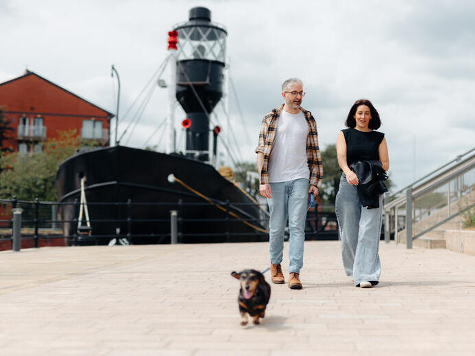 Two people walking a small dog along a paved waterfront with a historic ship and residential buildings in the background.