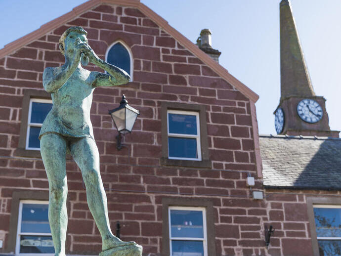 Bronze statue of a person blowing a horn in front of a red stone building and clock tower on a bright, clear day.
