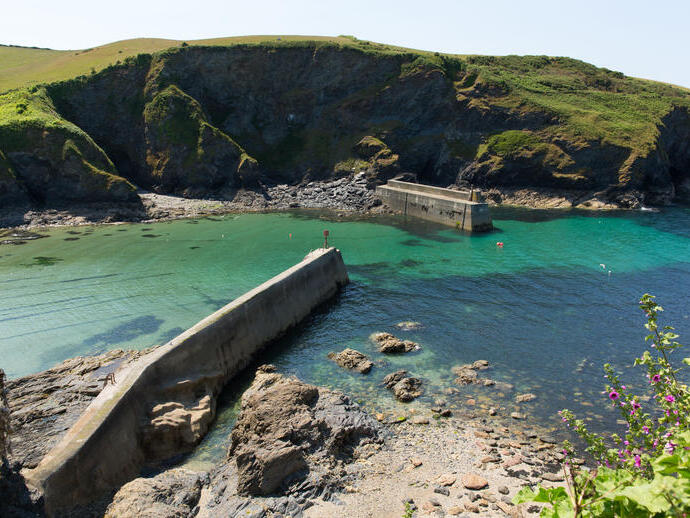 Clear turquoise waters, rocky shore, concrete harbor walls, and cliffs with green grass under a bright sky. Scenic coastal landscape.