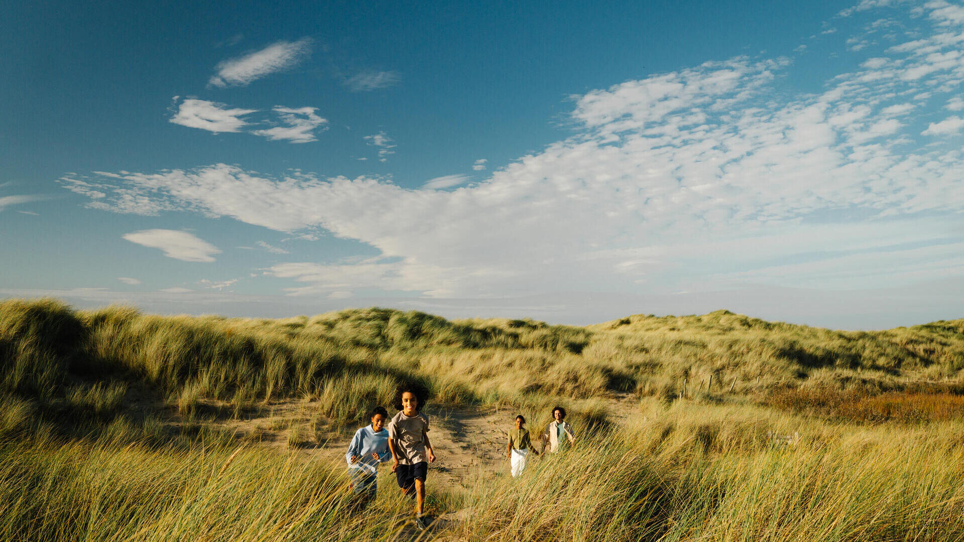 Children running through grassy sand dunes under a bright blue sky with scattered clouds.