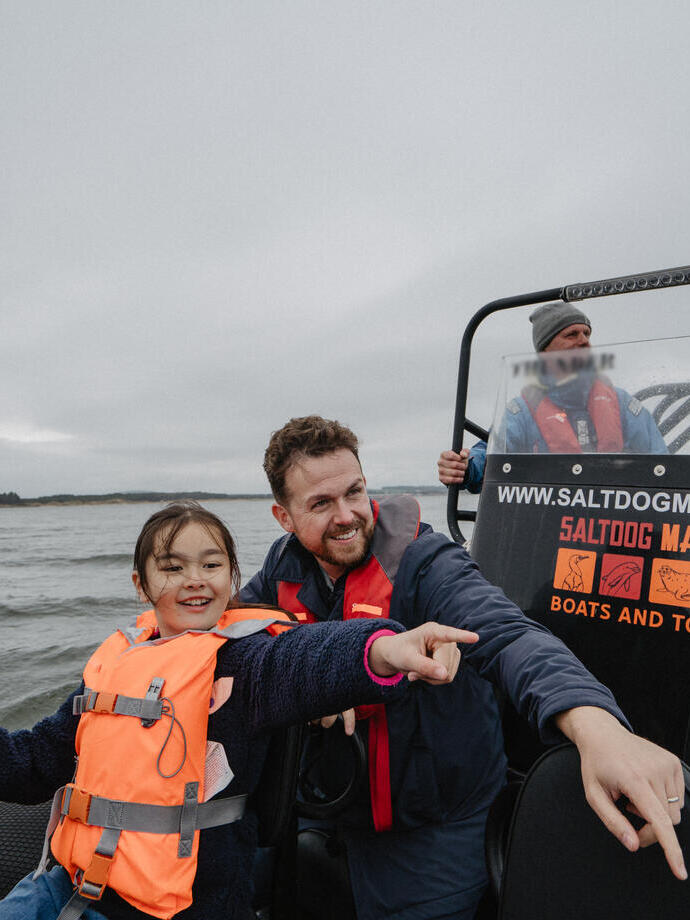 A man and a girl point at something from inside a boat with the Marine boat crew in the background.