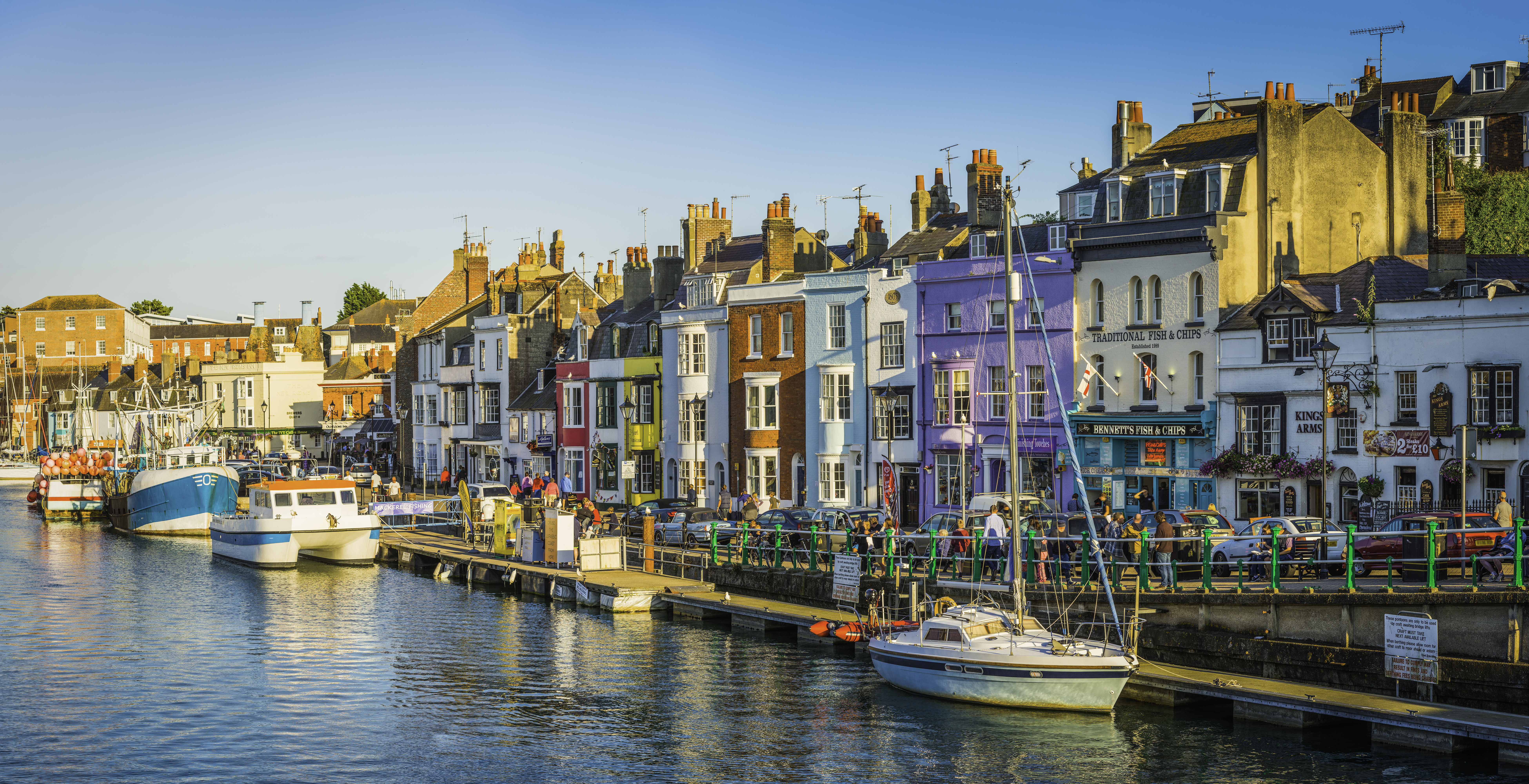 People walking up and down the water front of a busy seaside town lined with colourful terraced houses and boats anchored in the harbour