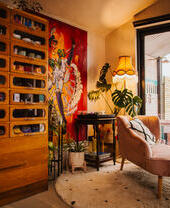 Une chaise longue rose pâle sur un tapis, donnant sur des baies vitrées. Une grande commode en bois à gauche et des œuvres d'art aux couleurs vives sur les murs.