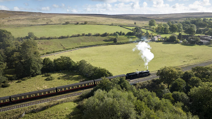A steam train travels through green countryside with fields, trees, stone walls, and distant farm buildings under a partly cloudy sky.
