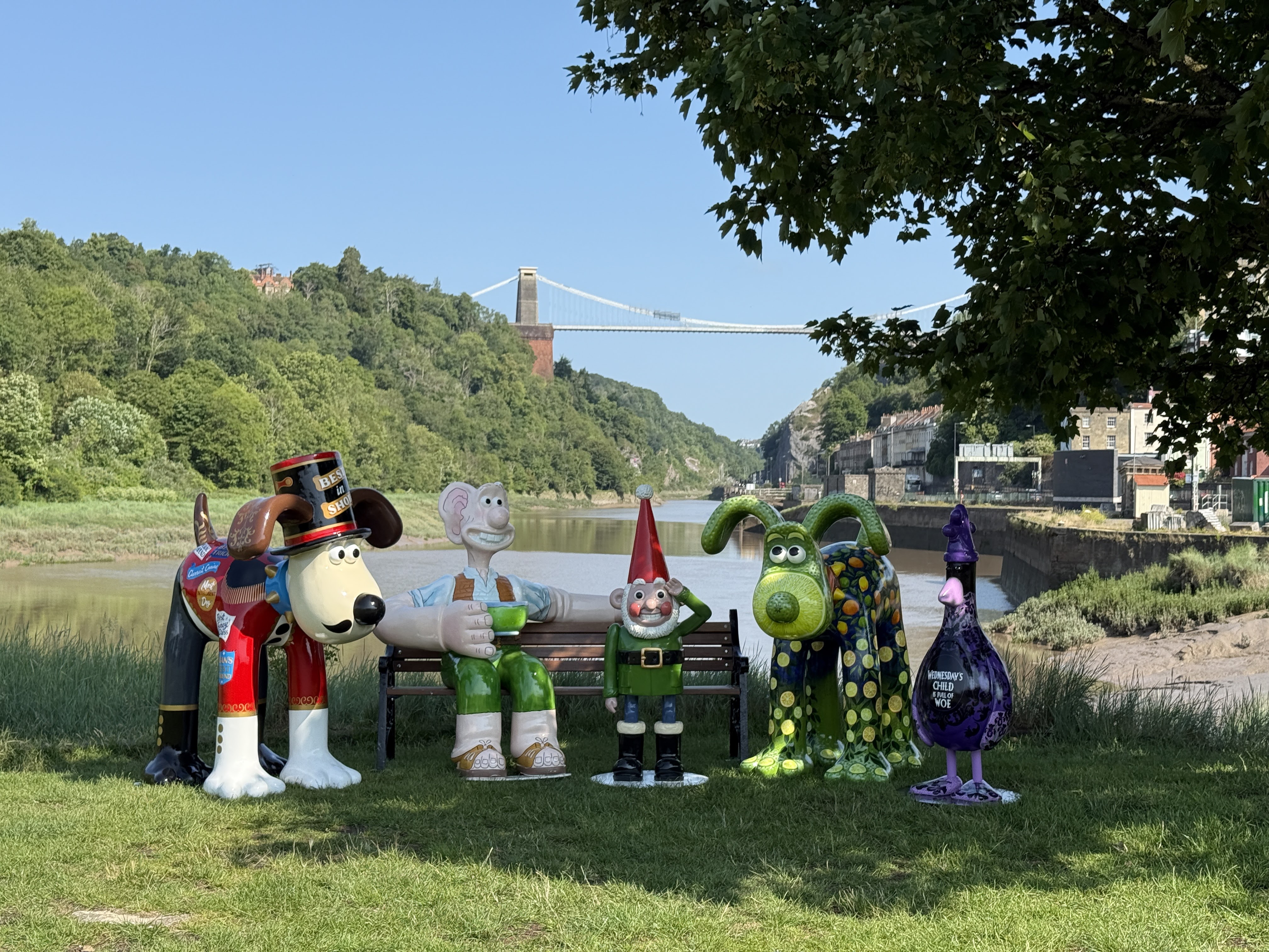 Sculptures of animated characters on grass near a river, with a suspension bridge and green hills in the background on a sunny day.