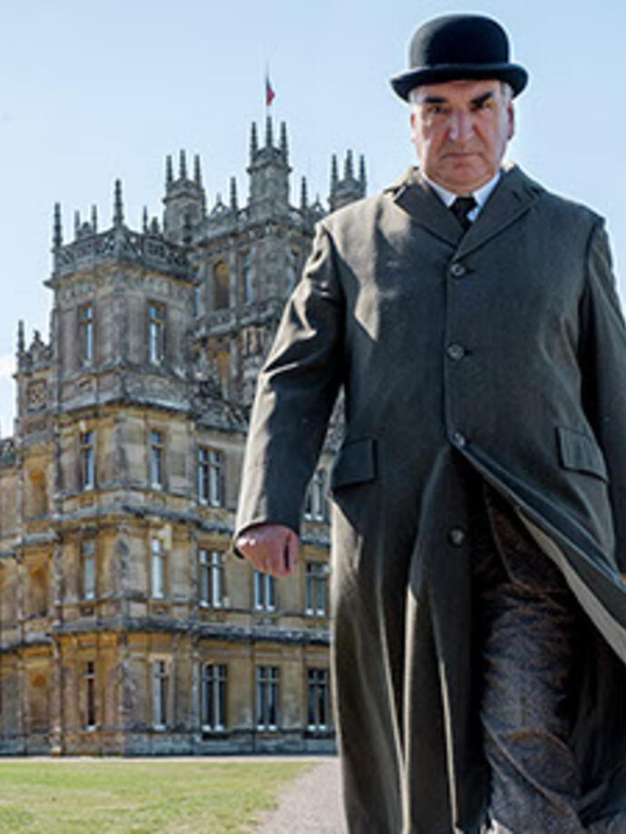 A person in period clothing walks in front of a grand historic mansion with ornate architecture and landscaped grounds.