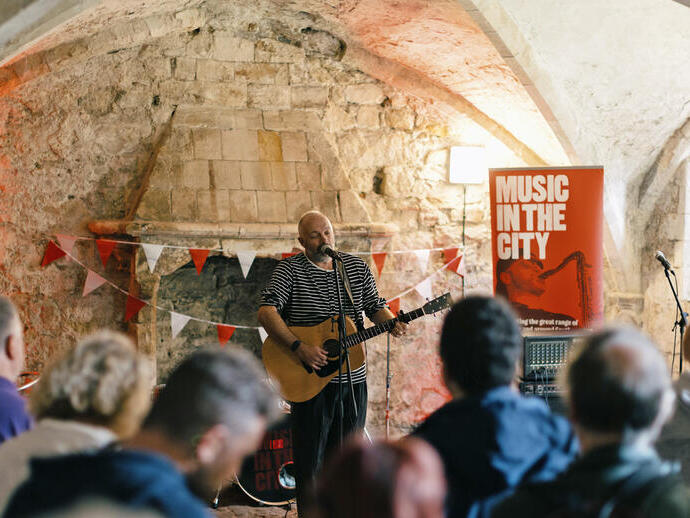 Musician performing with guitar in historic stone building, 'Music In The City' event banner and audience in foreground.