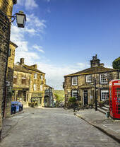 Stone street in a British village, historic stone buildings, red phone booth, blue sky, and a small café visible.