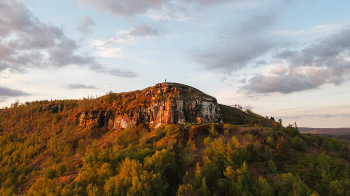 A lone person stands on a cliff top with forest below