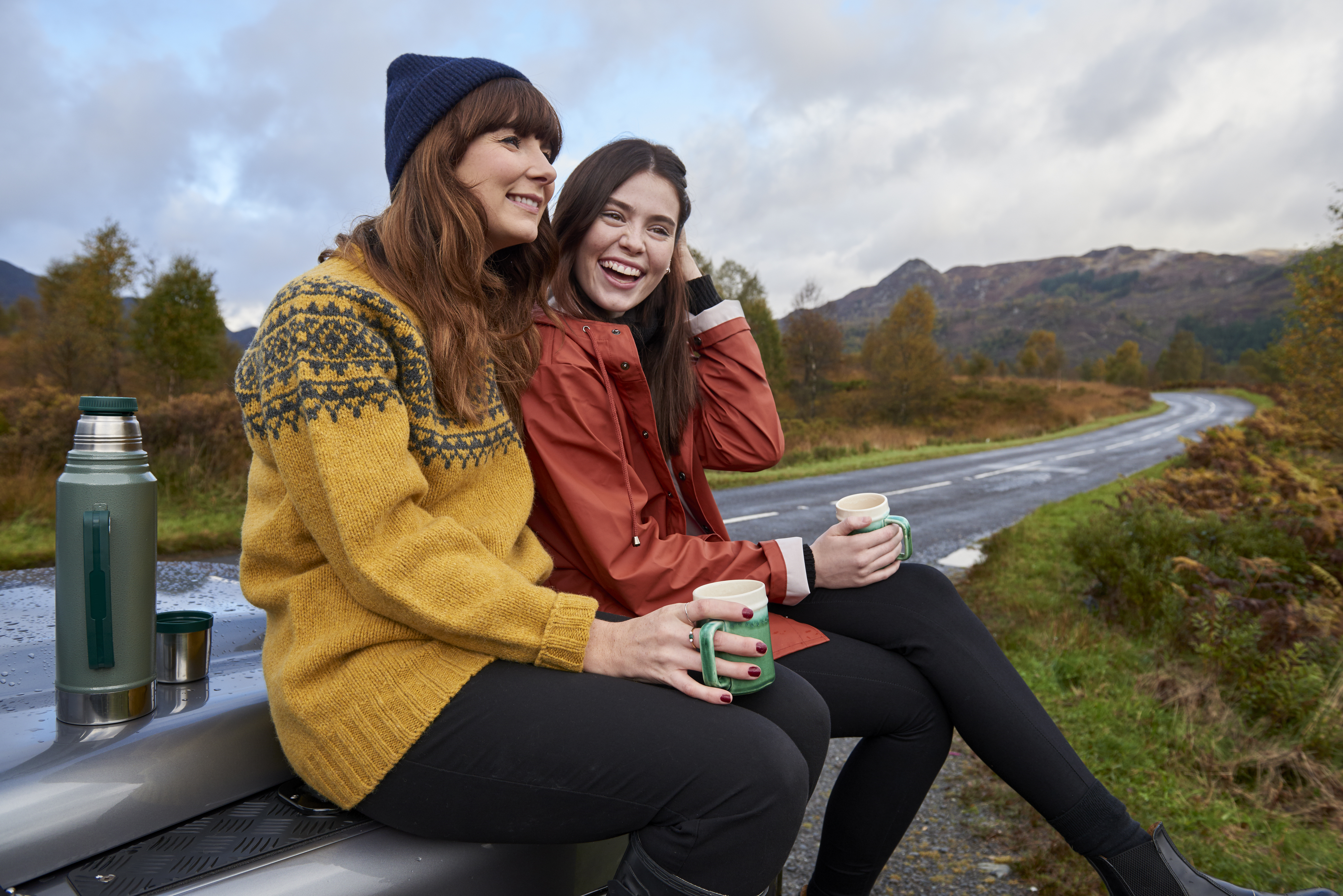Zwei Personen sitzen auf einer Motorhaube, trinken aus Tassen in ländlicher Umgebung, mit Thermoskanne daneben und einer kurvigen Straße im Hintergrund.